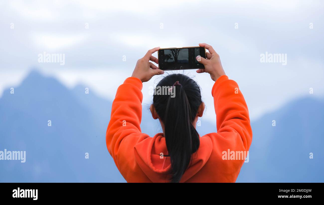 Rückansicht einer jungen Frau, die auf einem ruhigen Berg steht und am Telefon Fotos von der wunderschönen Berglandschaft bei Sonnenaufgang macht. Stockfoto