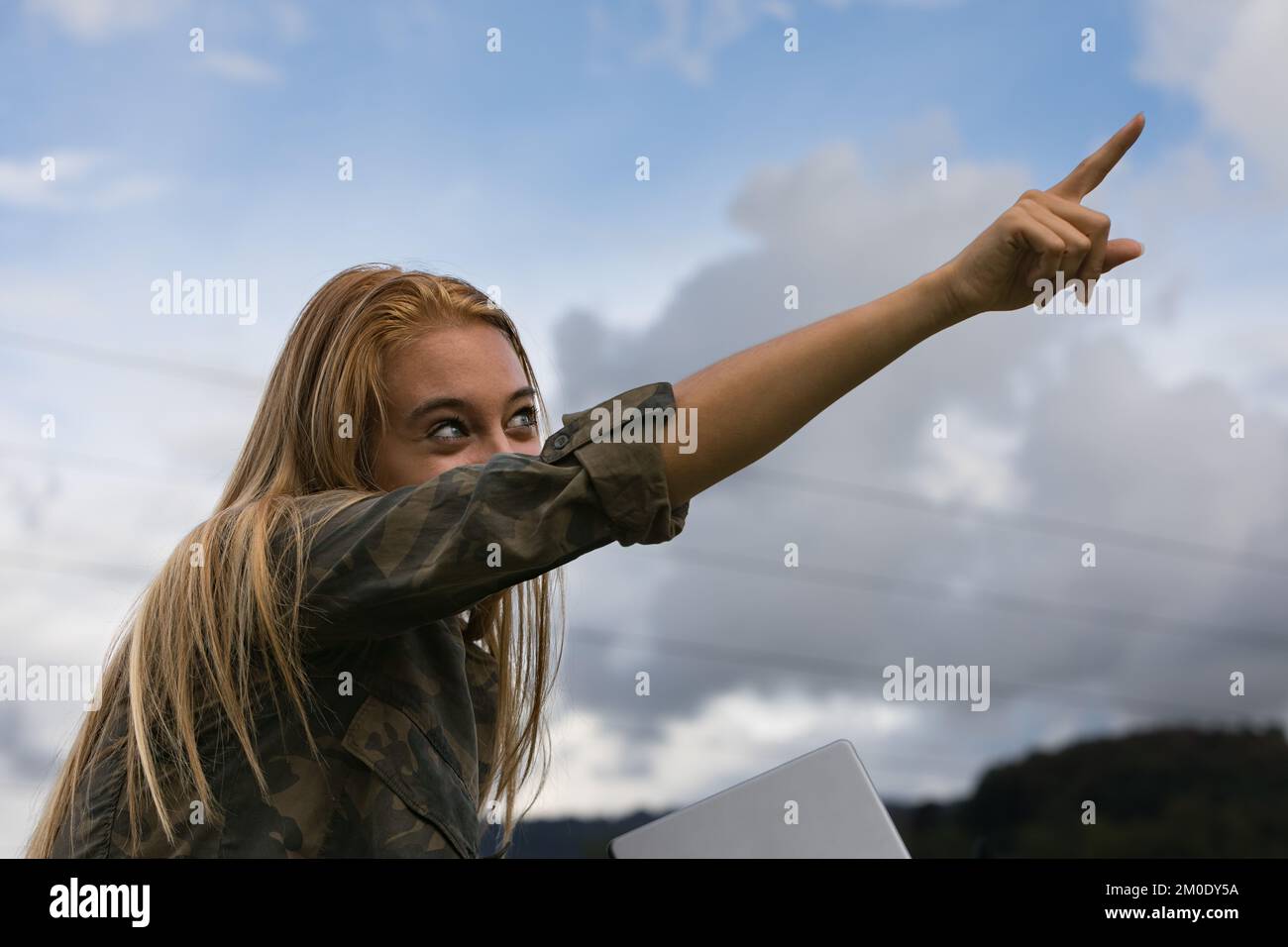 Der Blick auf ein androgynes Mädchen in einer jungen blonden Frau zeigt mit dem Zeigefinger zum Himmel, aber hinter ihr sind Hochspannungskabel, die elektrischen Strom tragen Stockfoto