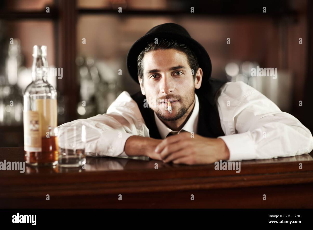 Alte Gewohnheiten sterben hart. Portrait eines gutaussehenden jungen Barkeepers, der neben ihm eine Flasche Whiskey raucht. Stockfoto