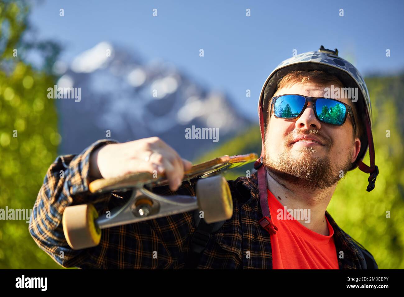 Porträt eines bärtigen Skaters mit Sonnenbrille und Helm, der sein Longboard im Hintergrund der Berge hält Stockfoto