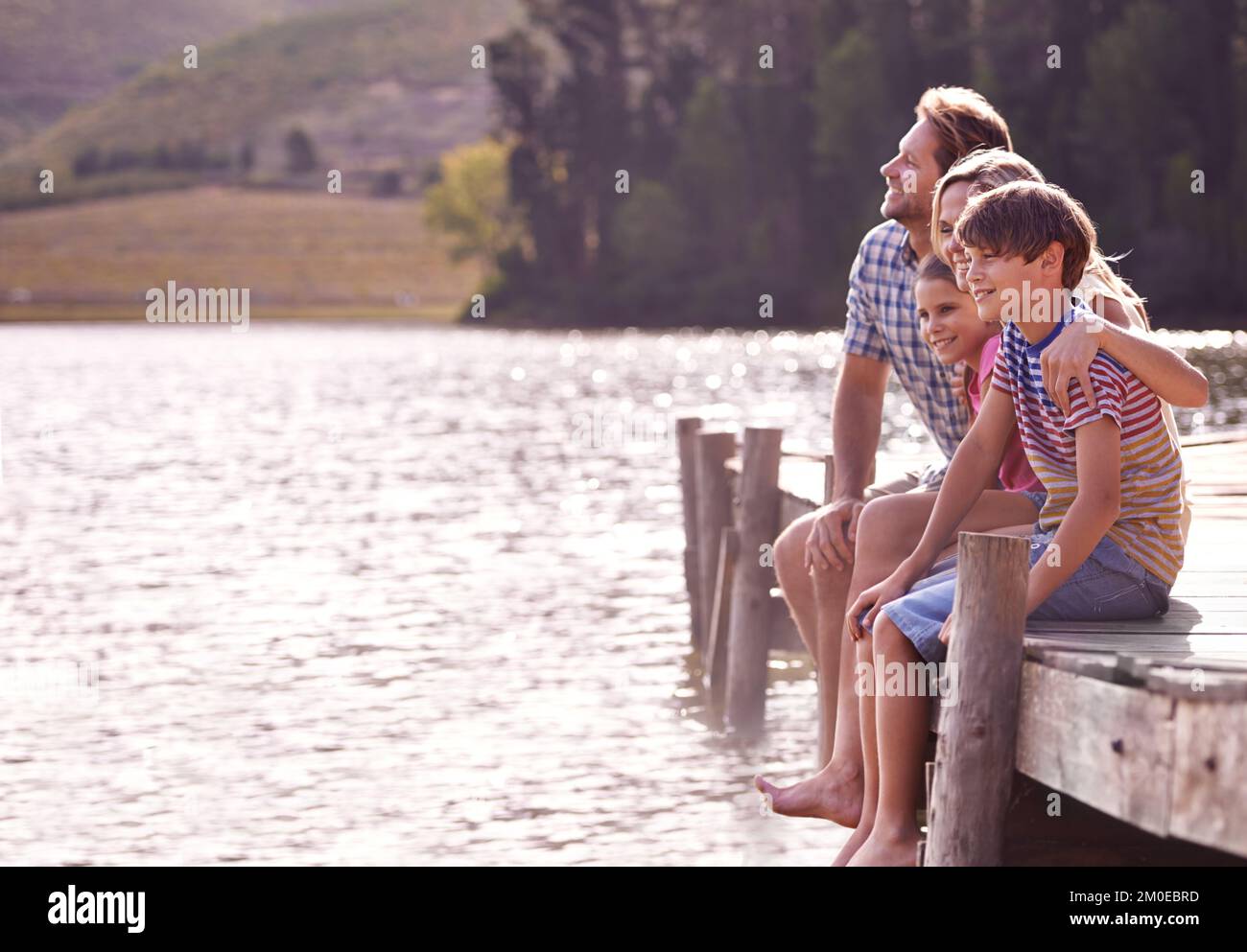 Die besonderen Momente der Zeit mit der Familie zu schätzen. Eine glückliche vierköpfige Familie, die auf einem Steg am See sitzt. Stockfoto