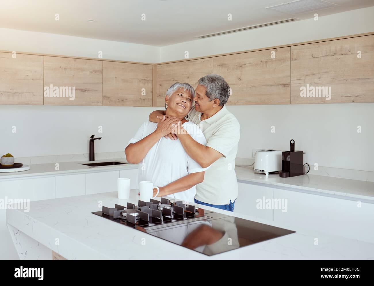 Gemischtes Seniorenpaar, das sich morgens zu Hause umarmt. Lächelnder älterer Ehemann und Ehefrau, die sich gegenseitig halten und in der Küche umarmen. Alles gute zum Ruhestand Stockfoto