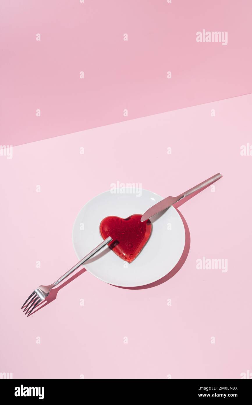 Herz aus rotem Gelee, silbernes Messer und Gabel auf weißem Teller und rosafarbenem Hintergrund. Creative Love oder Valentinstag Minimalkonzept. Stockfoto