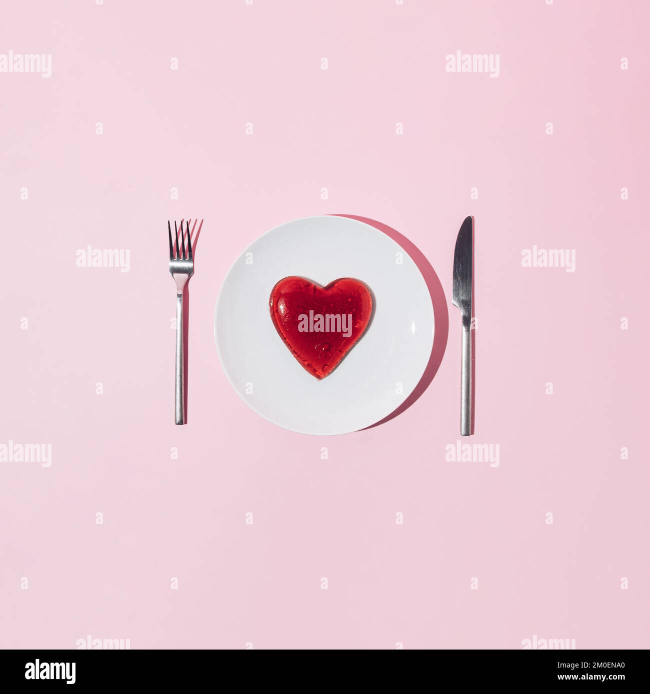 Herz aus rotem Gelee, silbernes Messer und Gabel auf weißem Teller und rosafarbenem Hintergrund. Liebe oder Valentinstag Minimalkonzept. Stockfoto