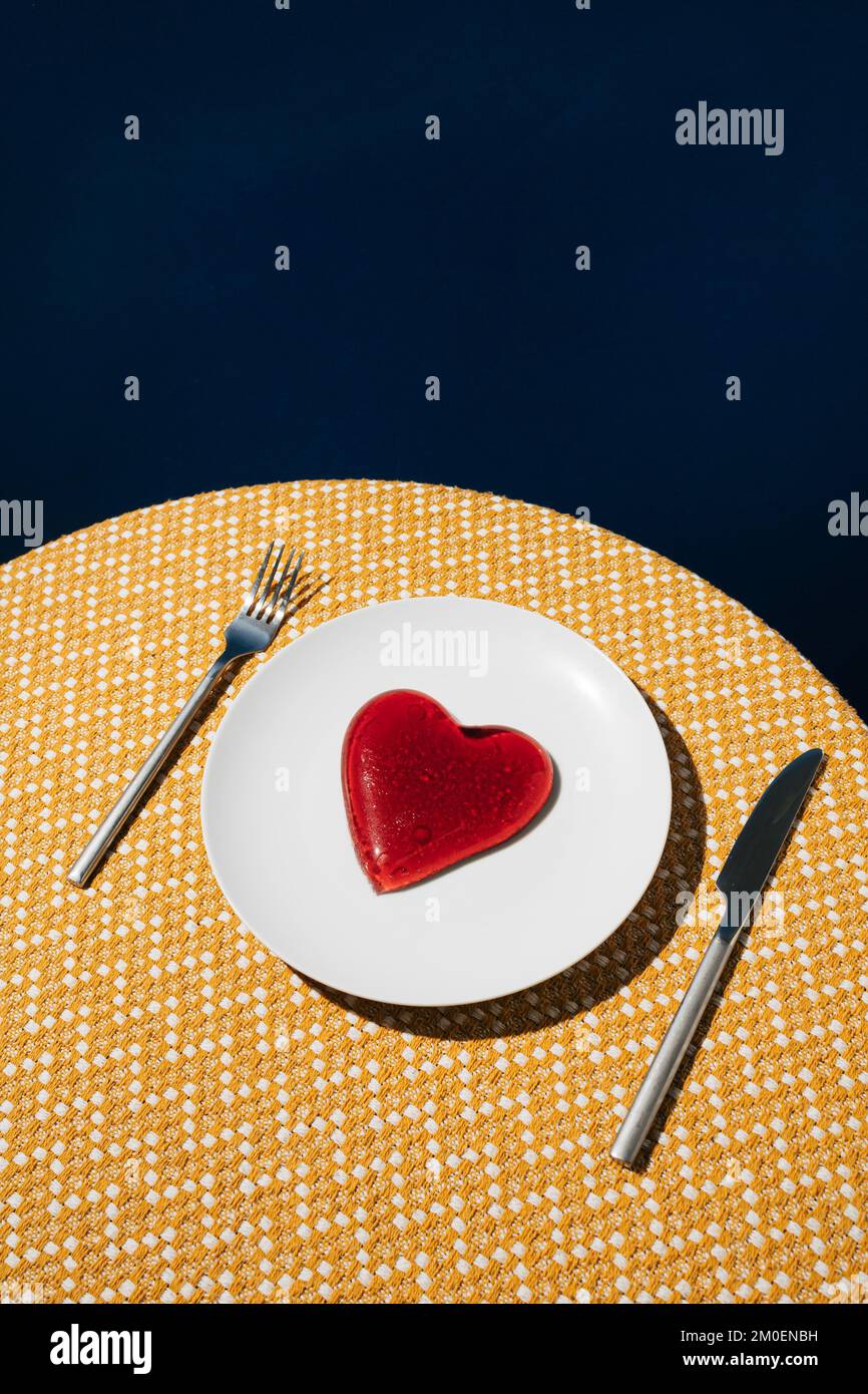 Rotes Gelee-Herz auf weißer Platte und gelbe Tischdecke mit dunkelblauem Hintergrund. Liebe oder Valentinstag Minimalkonzept. Stockfoto