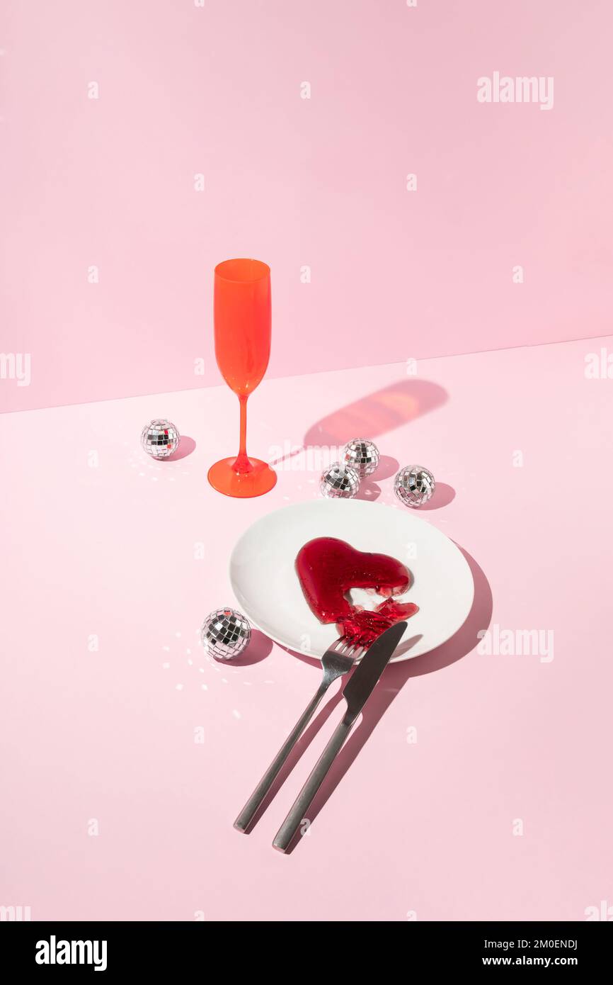 Rotes Gelee-Herz, Discokugeln, Messer und Gabel, Teller und Trinkglas auf pinkfarbenem Hintergrund. Liebe oder Valentinstag Minimalkonzept. Stockfoto