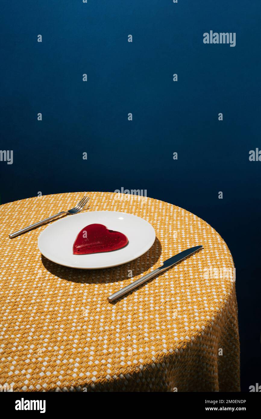 Rotes Gelee-Herz auf weißer Platte und gelbe Tischdecke mit dunkelblauem Hintergrund. Liebe oder Valentinstag Minimalkonzept. Stockfoto