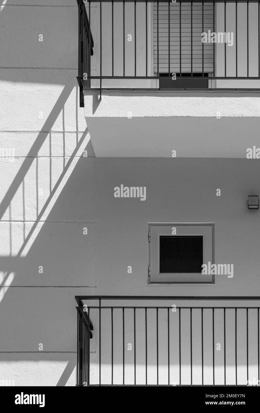 Weiße Fassade eines flachen Gebäudes mit minimalistischem Design und kleinen Fenstern. Der Schatten des schwarzen Geländers wird wie eine Treppe an die Wand projiziert Stockfoto