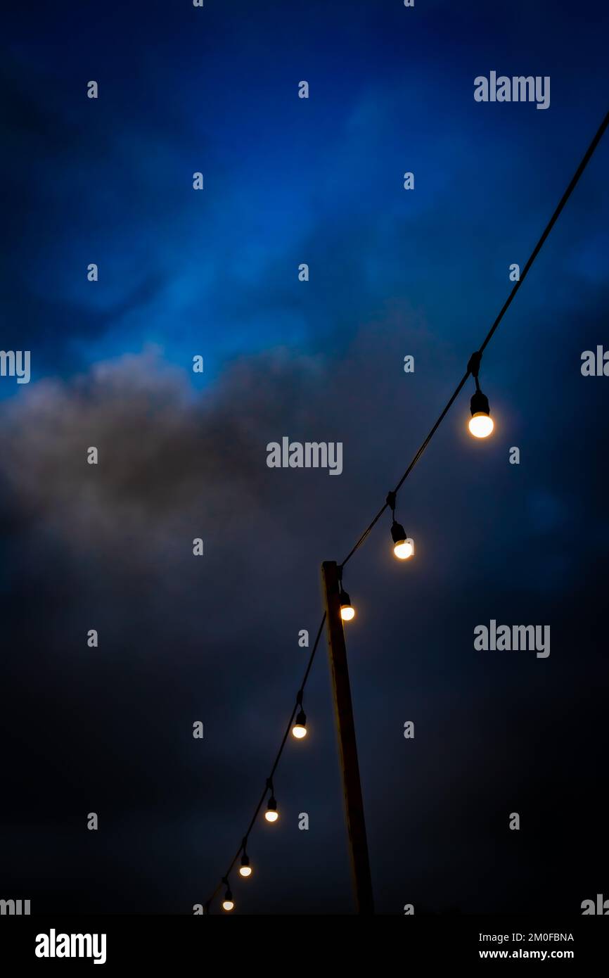 Eine Reihe von Glühbirnen, die von einem Holzpfosten gegen einen sehr dunklen und unheimlichen blauen Himmel gehalten werden. Viel Platz zum Kopieren. Ein gespenstisches und atmosphärisches Bild. Stockfoto