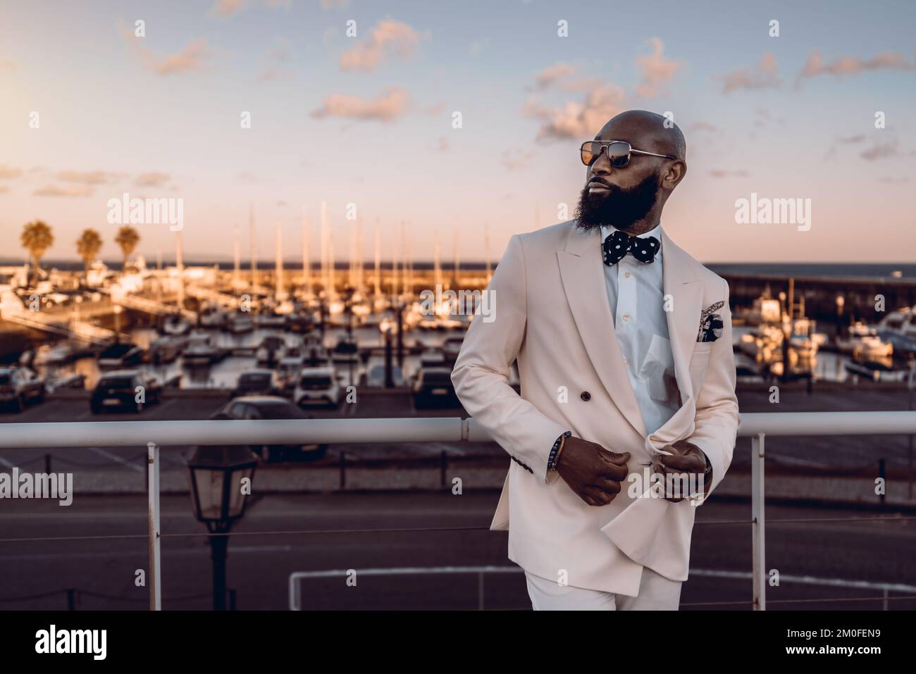 Ein stilvoller bärtiger schwarzer Mann posiert zur goldenen Stunde; der afrikanische Mann trägt einen beigen Anzug mit blauer Fliege und weißen Punkten sowie einer Sonnenbrille Stockfoto