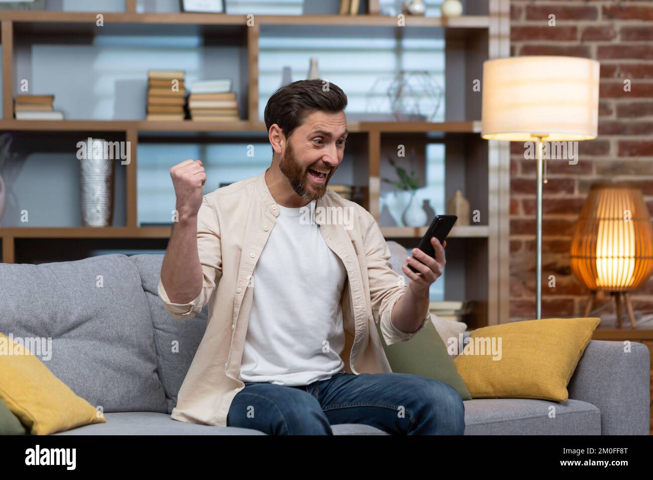 Mann zu Hause auf dem Sofa mit Smartphone, feiert den Sieg im Wohnzimmer und liest Happy-News-Benachrichtigungen vom Telefon online, hält Hand-hoch-Geste des Triumphes und Erfolg sitzt auf dem Sofa. Stockfoto