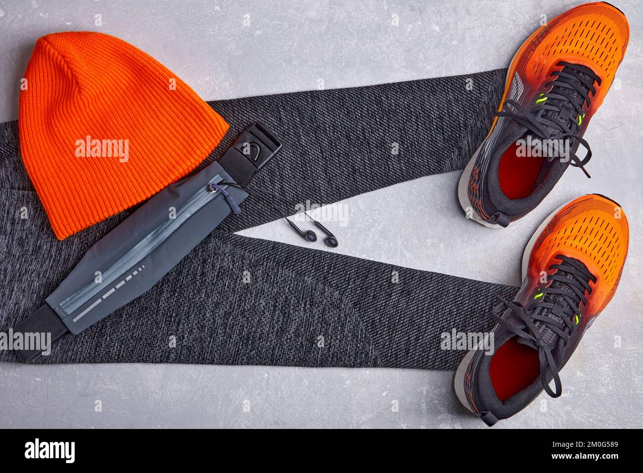 Eine orangefarbene Strickmütze, warme Sportleggings, eine gürteltasche mit Kopfhörern und orangefarbenen Turnschuhen auf hellem Betonhintergrund Stockfoto