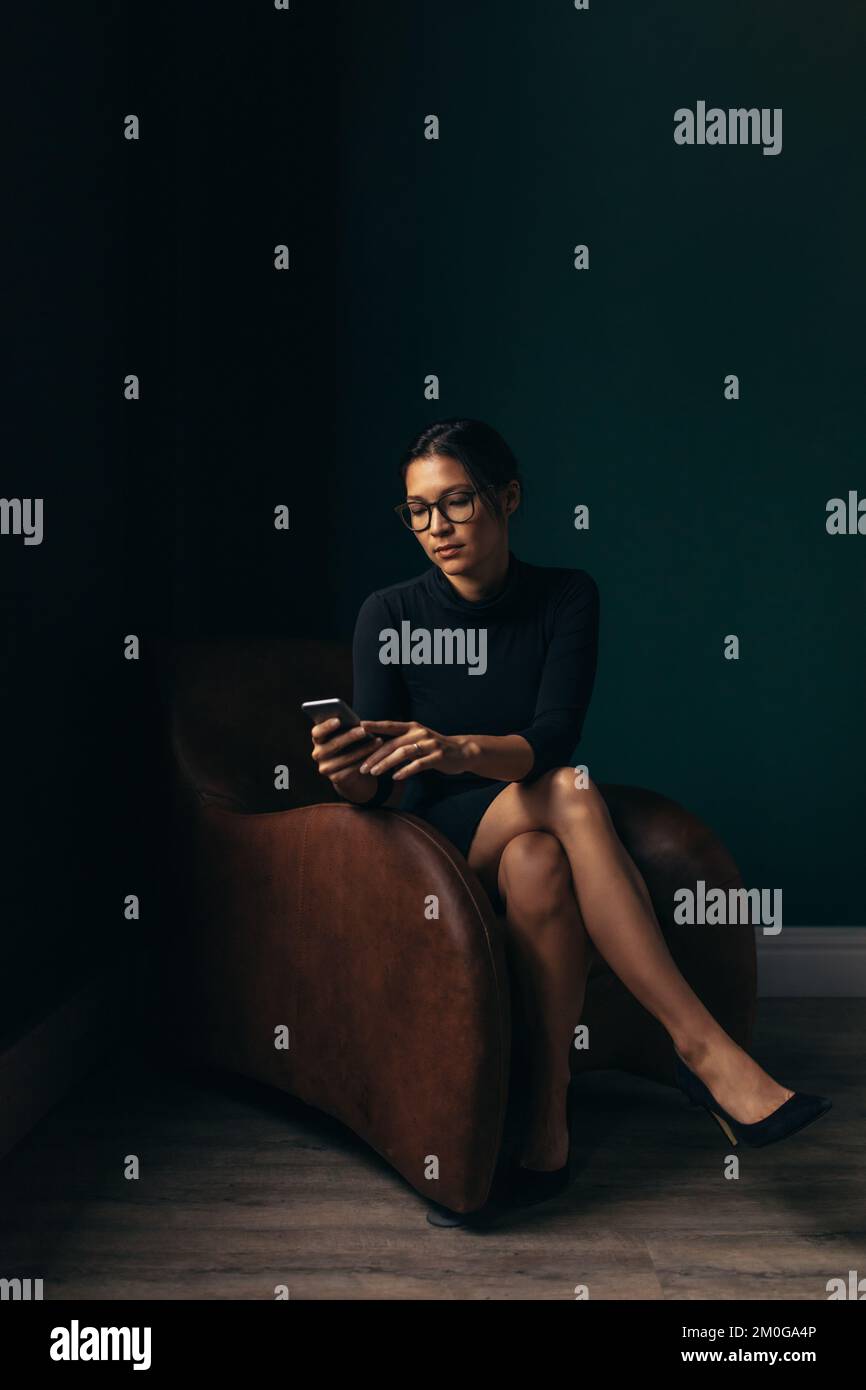 Eine anspruchsvolle Frau, die ein Mobiltelefon benutzt, während sie in ihrem Büro auf einem Sessel sitzt. Eine moderne Frau, die eine SMS auf ihrem Smartphone liest. Stockfoto
