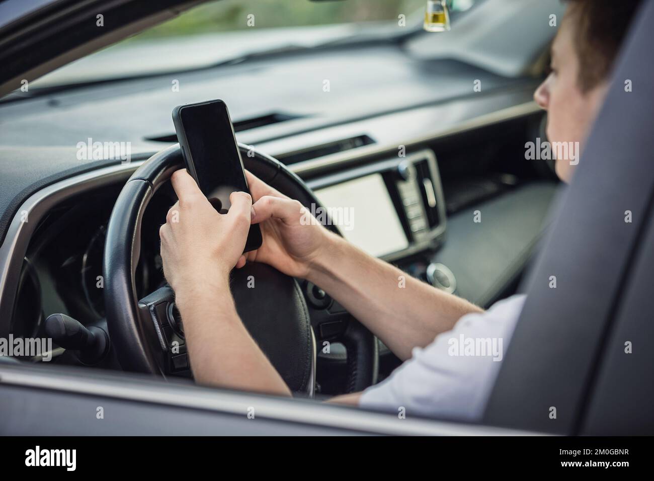Ein junger Fahrer, der sich vor dem Lenkrad von seinem Telefon abgelenkt hat und sein Smartphone während der Fahrt mit einer Hand benutzte. Risiko- und Gefahrensituation Stockfoto
