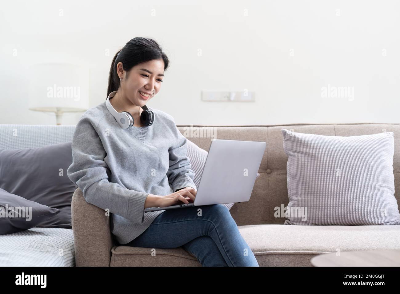 Foto einer jungen asiatischen Frau, die fröhlich lächelt und auf der Couch sitzt, um im Haus nach einem Laptop zu suchen Stockfoto