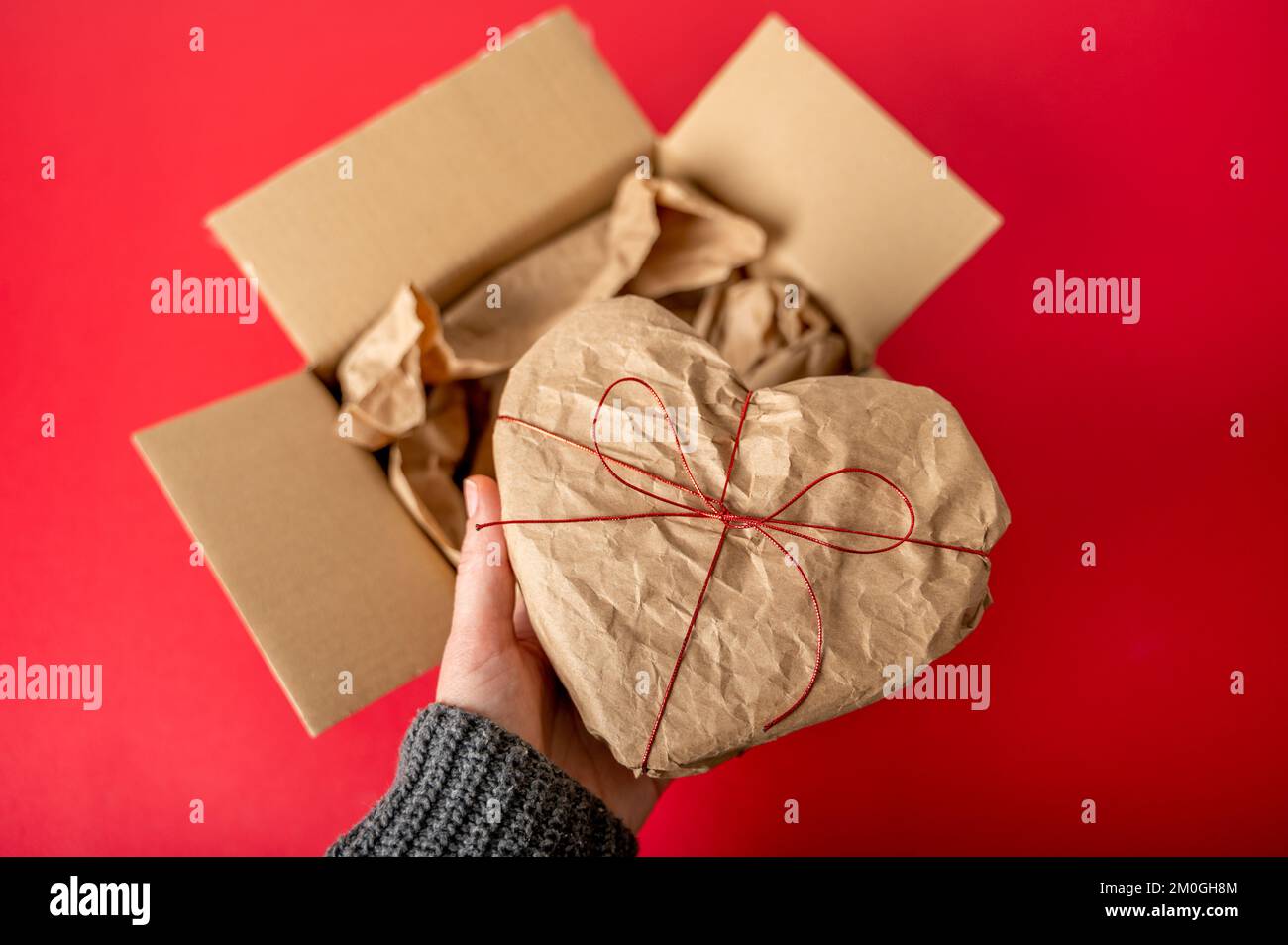 Hände in herzförmiger Geschenkbox, verpackt in Kraftpapier auf rotem Hintergrund. Ich habe nur das Paket ausgepackt Stockfoto