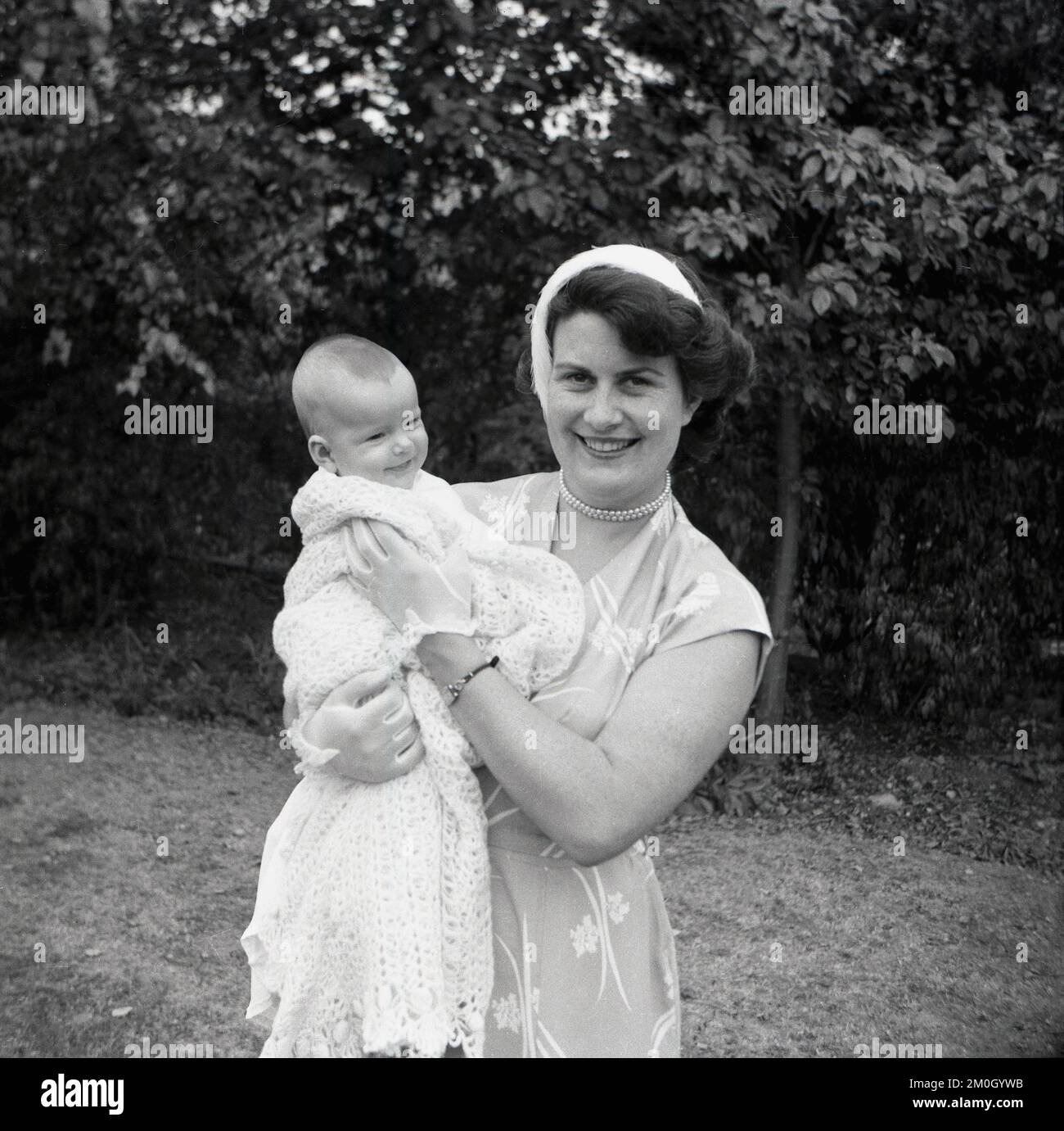 1950er, historisch, taufend, draußen für ein Foto zu stehen, eine glückliche Mutter hält ihr gerade tauftes Kind, das in einen Babyschal mit Häkelmuster eingewickelt ist, mit einem süßen Lächeln auf seinem kleinen Gesicht, England, Großbritannien. Stockfoto