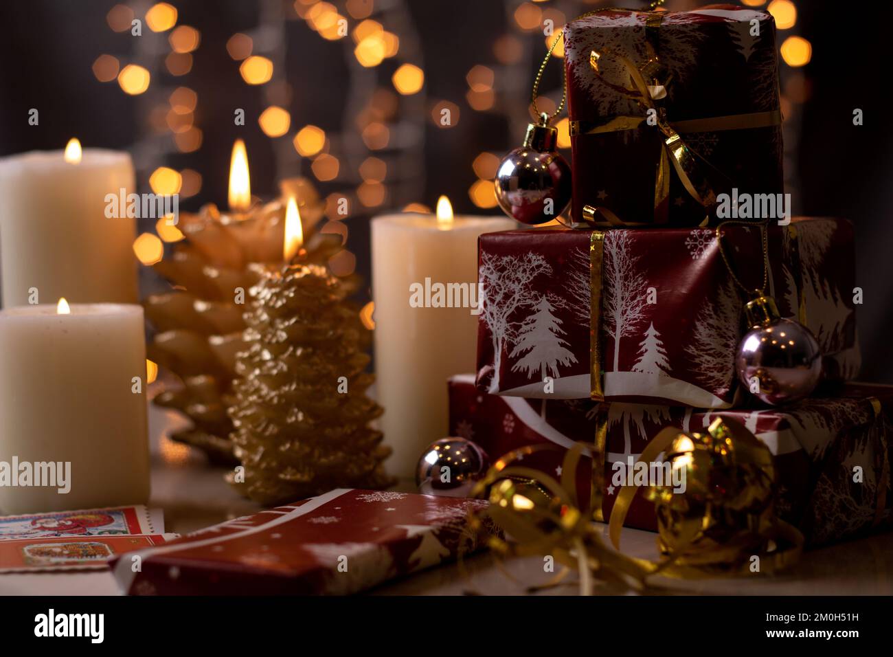 Fotogeschenke in rotem Papier stehen aufrecht in einer Reihe neben beleuchteten Kerzen auf dem Hintergrund mit einem Bokeh aus Weihnachtslichtern Stockfoto