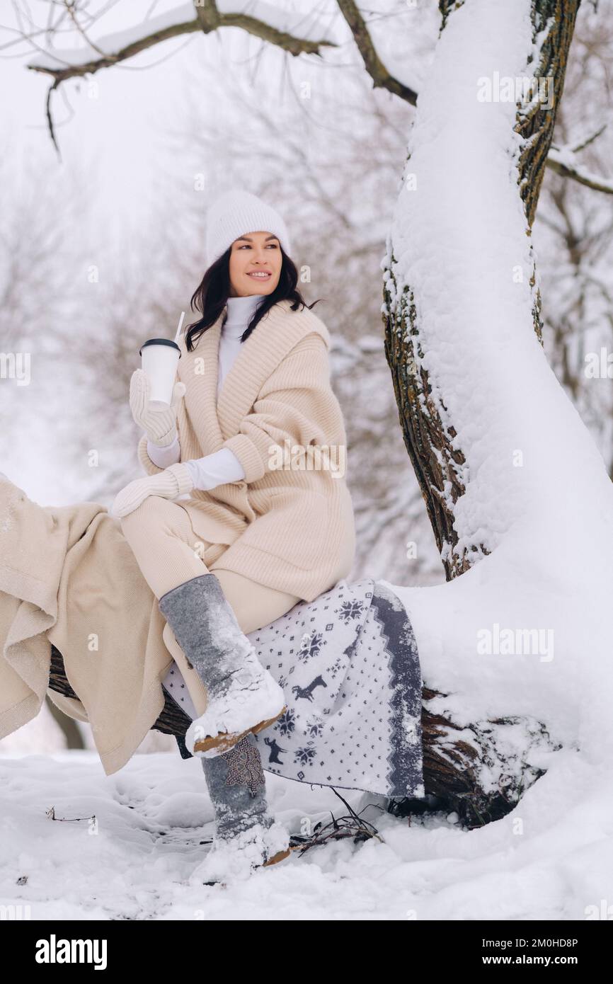 Ein hübsches Mädchen mit einer beigen Strickjacke und einem weißen Hut, das Tee in einem verschneiten Winterwald in der Nähe eines Sees trinkt Stockfoto