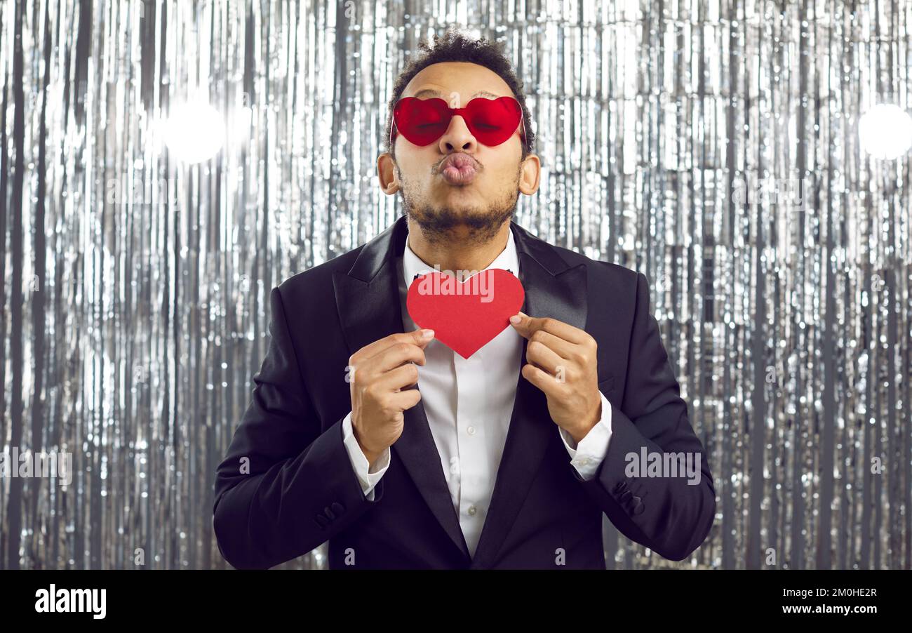 Lustiger Mann in Anzug und Herzbrille, der Valentinskarte hält und seine Lippen für einen Kuss schmollt Stockfoto