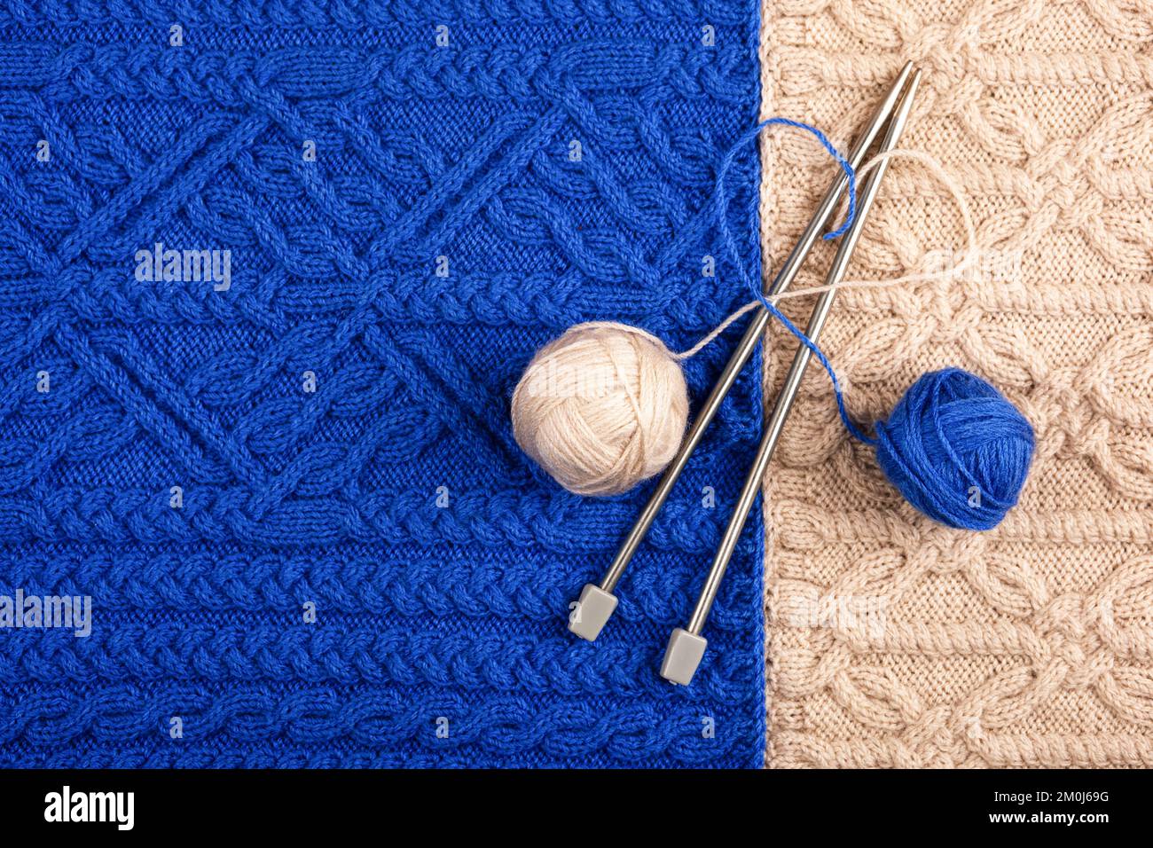 Zwei Fadenbälle und Stricknadeln liegen auf einem blau-beigen Strickstoff. Das Konzept, Zeit zu Hause zu verbringen und warme Kleidung für wi zu stricken Stockfoto