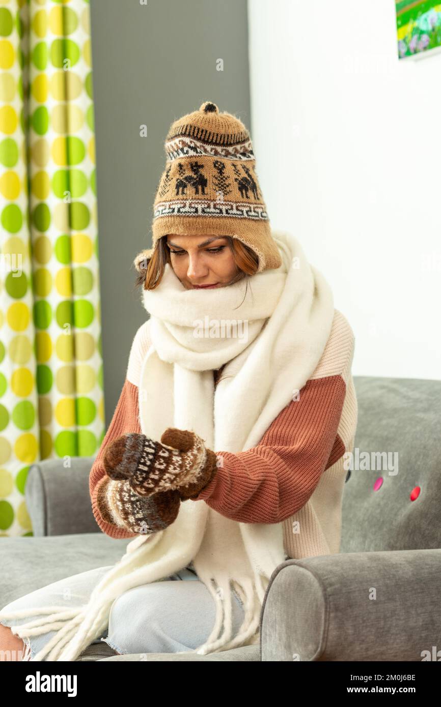 Eine junge Frau, die auf dem Sofa sitzt, einen Wollmütze, einen Schal trägt und Wollhandschuhe anzieht Stockfoto