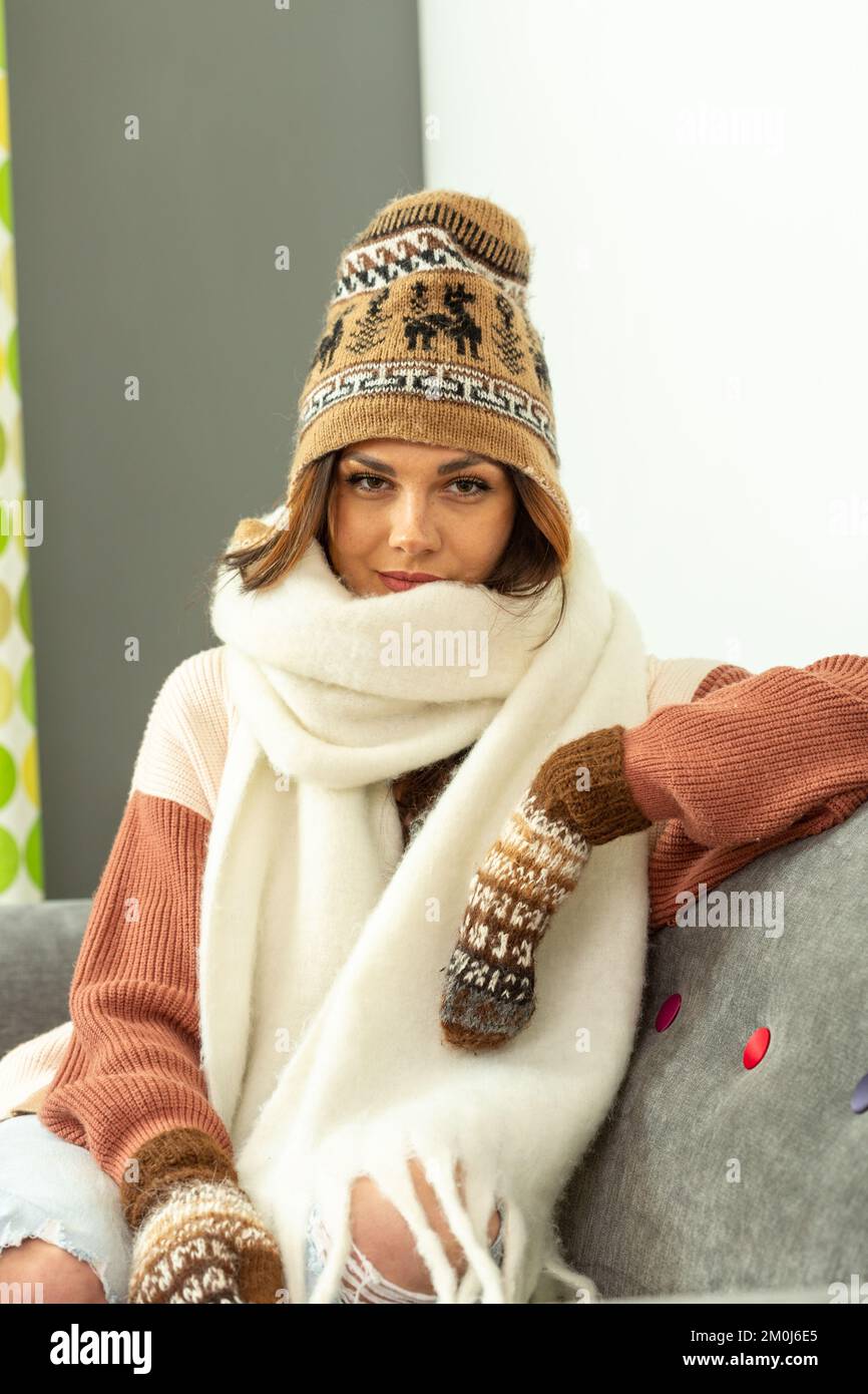 Eine junge Frau, die auf dem Sofa sitzt und einen Wollmütze, Schal und Fäustlinge trägt Stockfoto