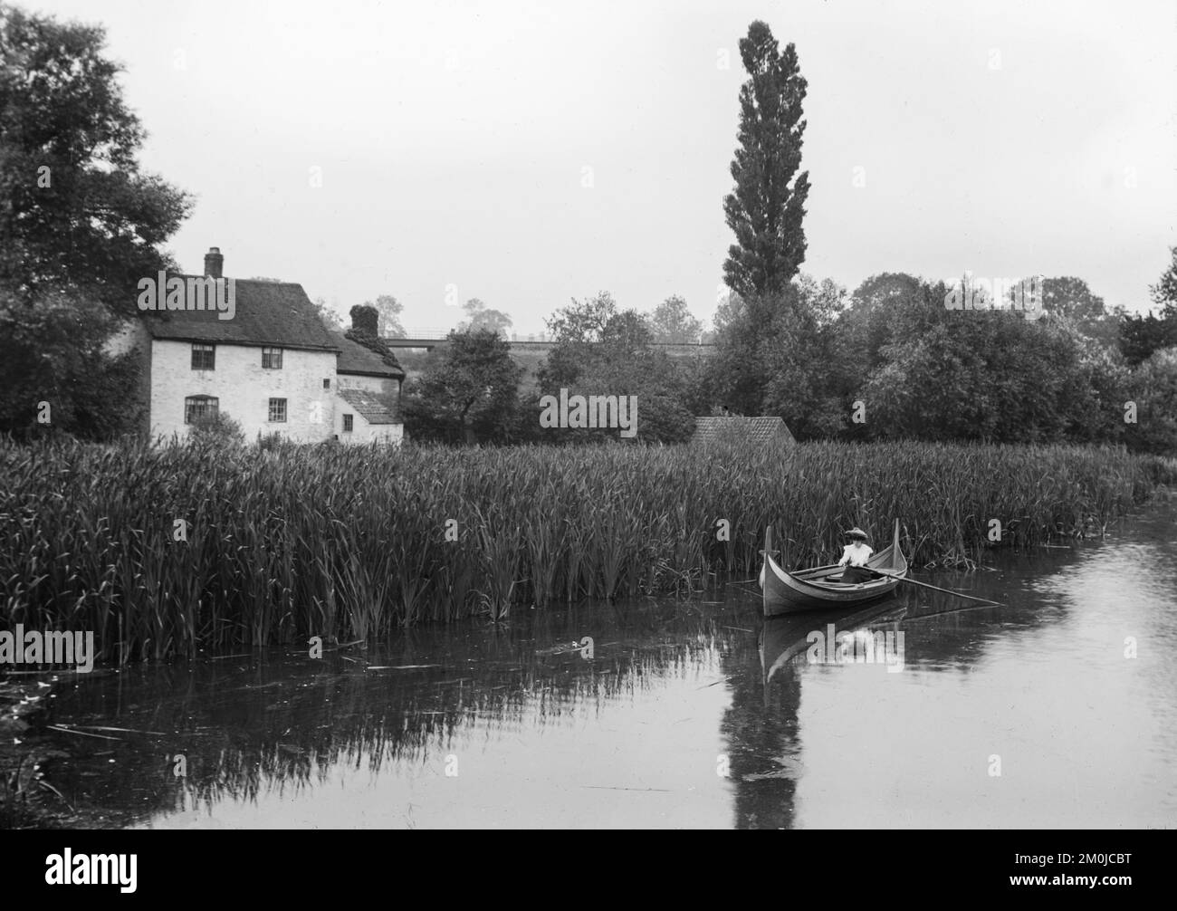Schwarzweißes englisches Foto aus dem späten 19.. Jahrhundert, das eine Frau in einem Ruderboot auf einem Fluss zeigt, neben einigen großen Anlässen mit einem Haus oder einer Hütte in der Nähe. Stockfoto