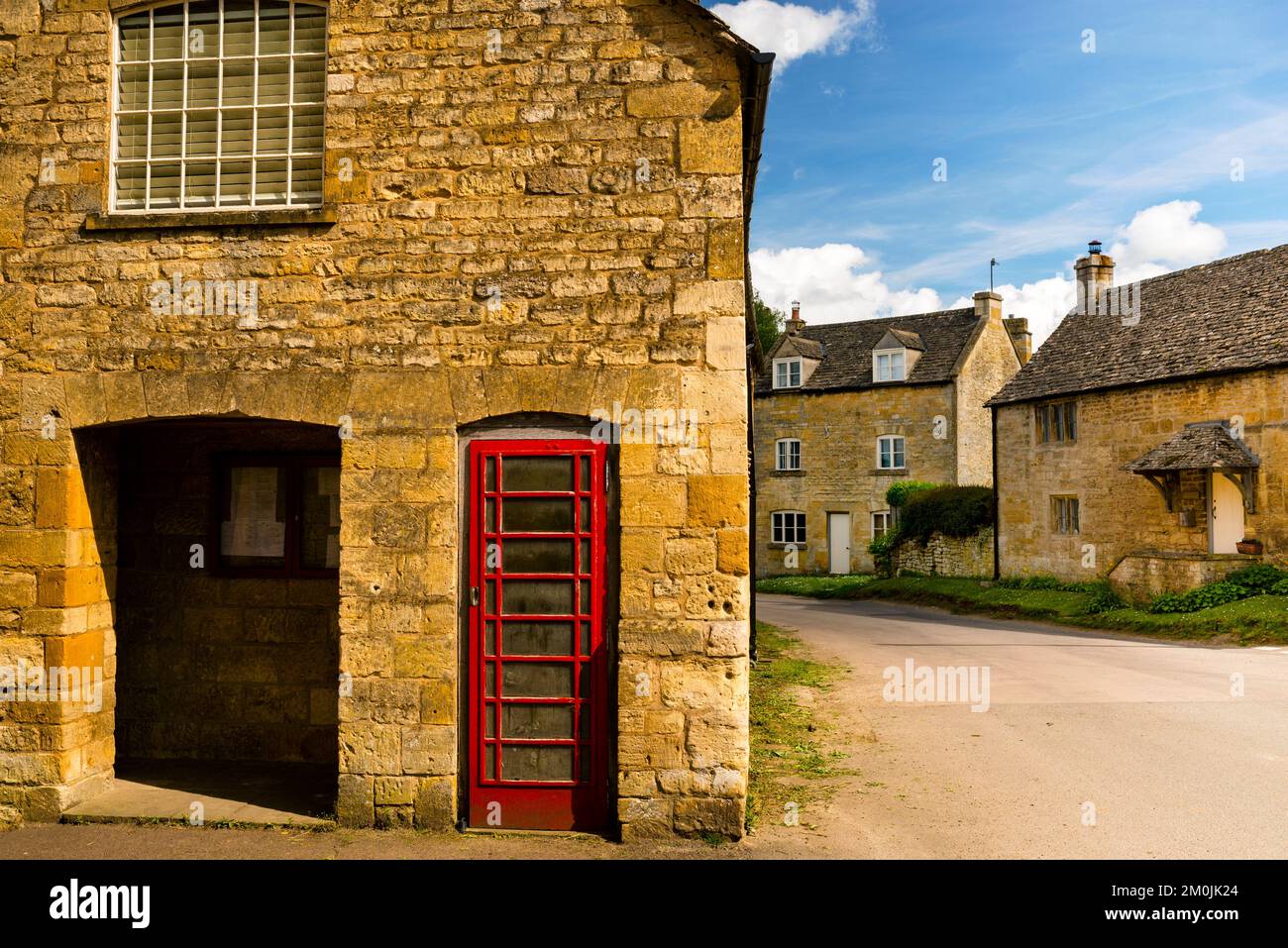 Guiting Powers rote Telefonzelle versteckt in goldenem gelben Cotswolds-Stein, England. Stockfoto