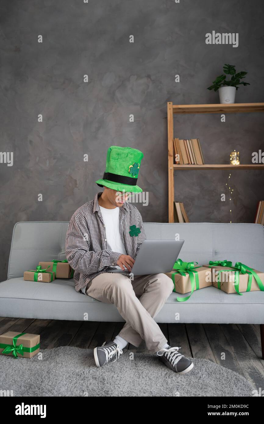 Das Online-Shopping-Konzept des St. Patricks Day. Männlicher Käufer mit Laptop auf der Couch. Der Typ kauft Geschenke, bereitet sich auf den St. Patricks-Tag vor, sitzt unter Geschenkschachteln. Sp Stockfoto