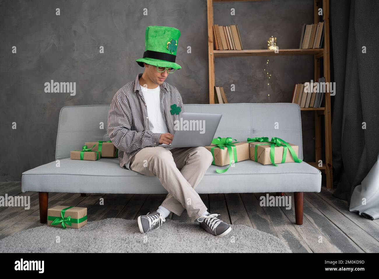 Das Online-Shopping-Konzept des St. Patricks Day. Männlicher Käufer mit Laptop auf der Couch. Der Typ kauft Geschenke, bereitet sich auf den St. Patricks-Tag vor, sitzt unter Geschenkschachteln. Sp Stockfoto