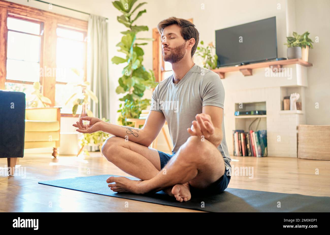 Ein junger kaukasischer Mann, der mit gekreuzten Beinen und geschlossenen Augen sitzt, meditiert in Harmonie mit einer Fingerbewegung, während er zu Hause Yoga praktiziert. Ruhig Stockfoto