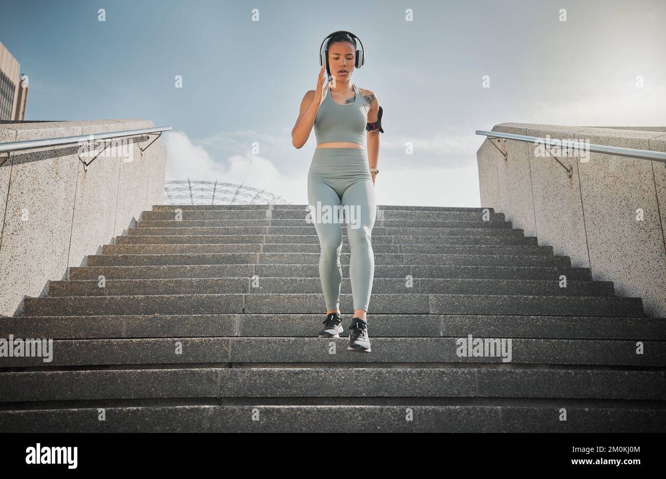 Junge gemischte Sportlerin hört Musik über Kopfhörer, während sie draußen die Treppen eines Gebäudes hinunterläuft. Junge Frau konzentrierte sich auf ihre Geschwindigkeit Stockfoto