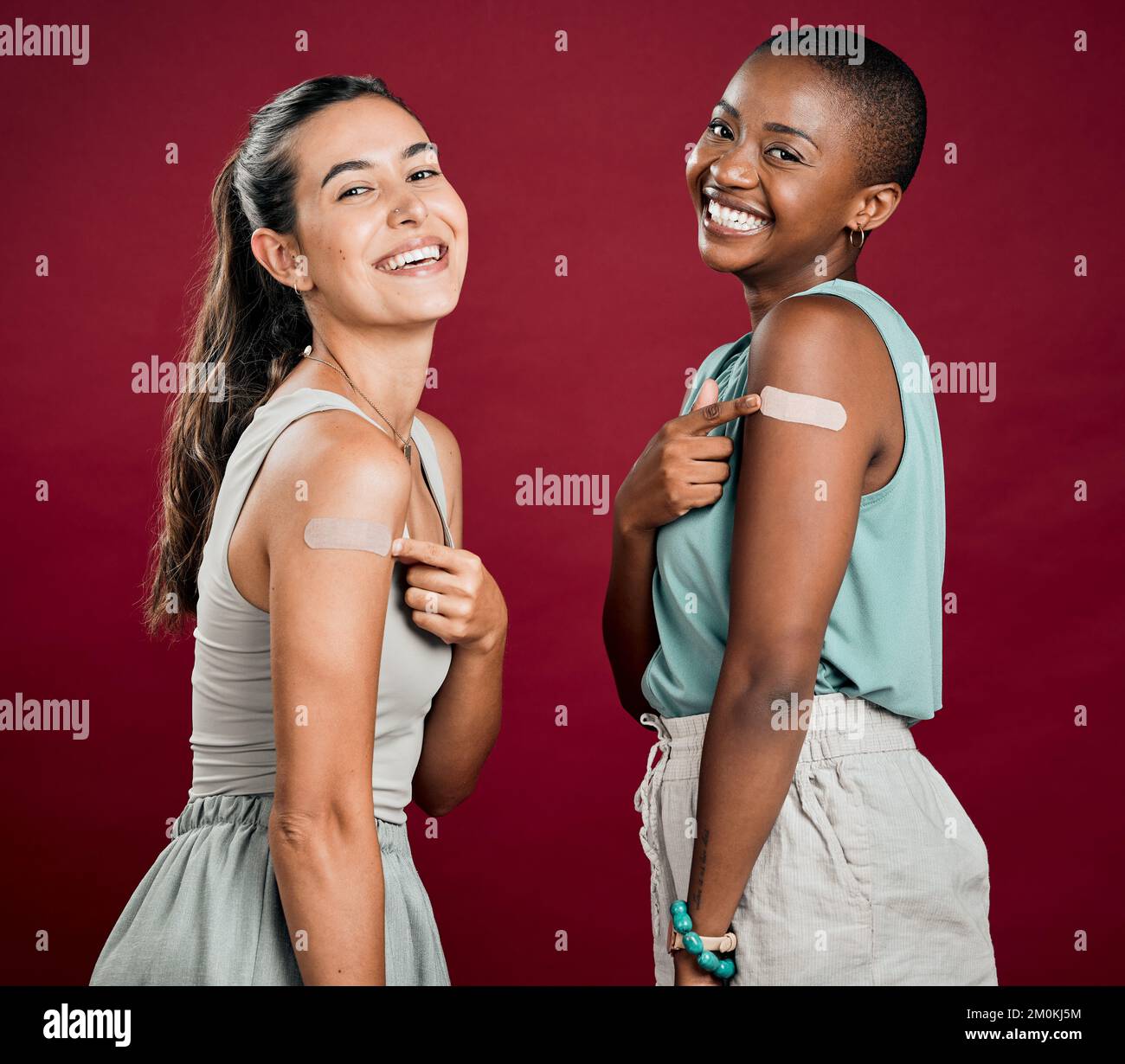Covid-geimpfte afroamerikanische und gemischte Rassen Frauen zeigen auf Armgips. Zwei glückliche Menschen, isoliert auf rotem Studiohintergrund Stockfoto