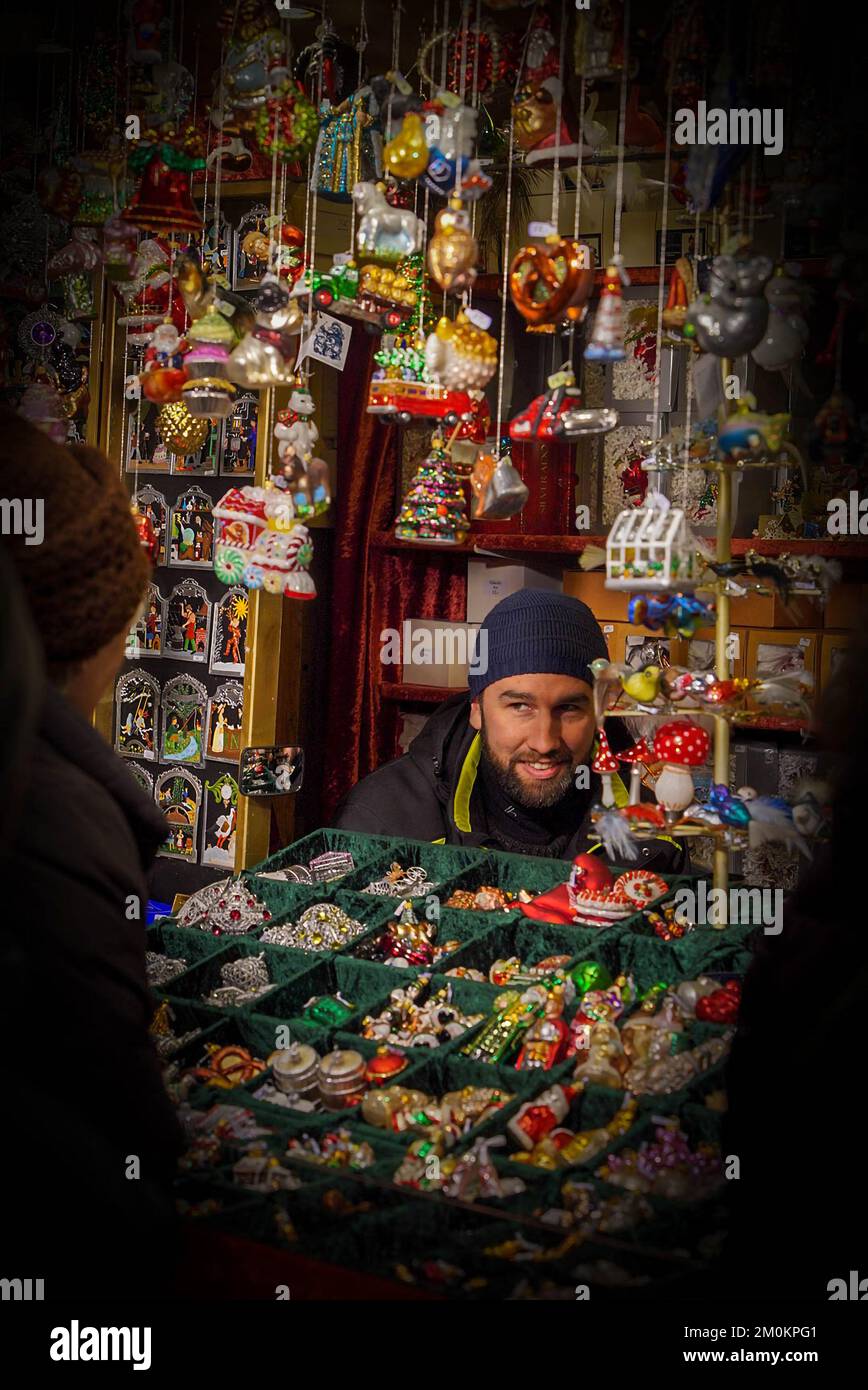 Ein männlicher Verkäufer sitzt nachts auf einem Weihnachtsmarkt in München am Fenster einer Theke mit Silvesterspielzeug gerahmt Stockfoto
