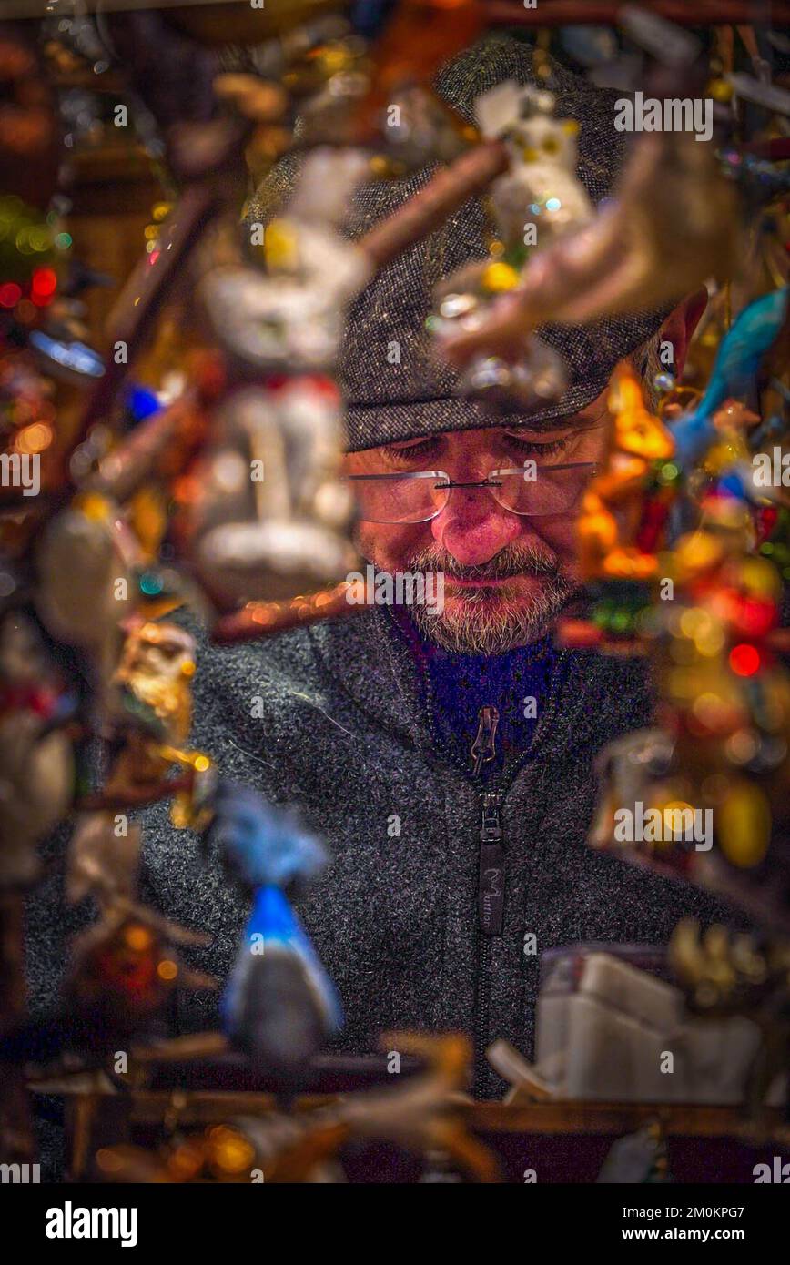 Ein männlicher Verkäufer sitzt nachts auf einem Weihnachtsmarkt in München am Fenster einer Theke mit Silvesterspielzeug gerahmt Stockfoto
