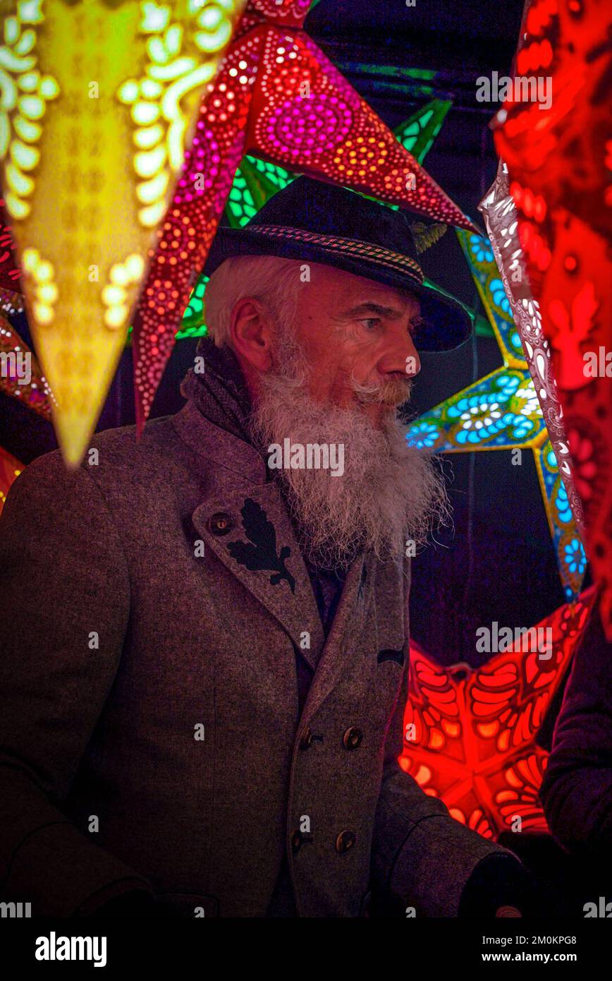 Ein erwachsener Mann mit grauem Bart bleibt unter den beleuchteten dekorativen Sternen an einer Theke mit Silvesterspielzeug auf einem Weihnachtsmarkt in München Stockfoto