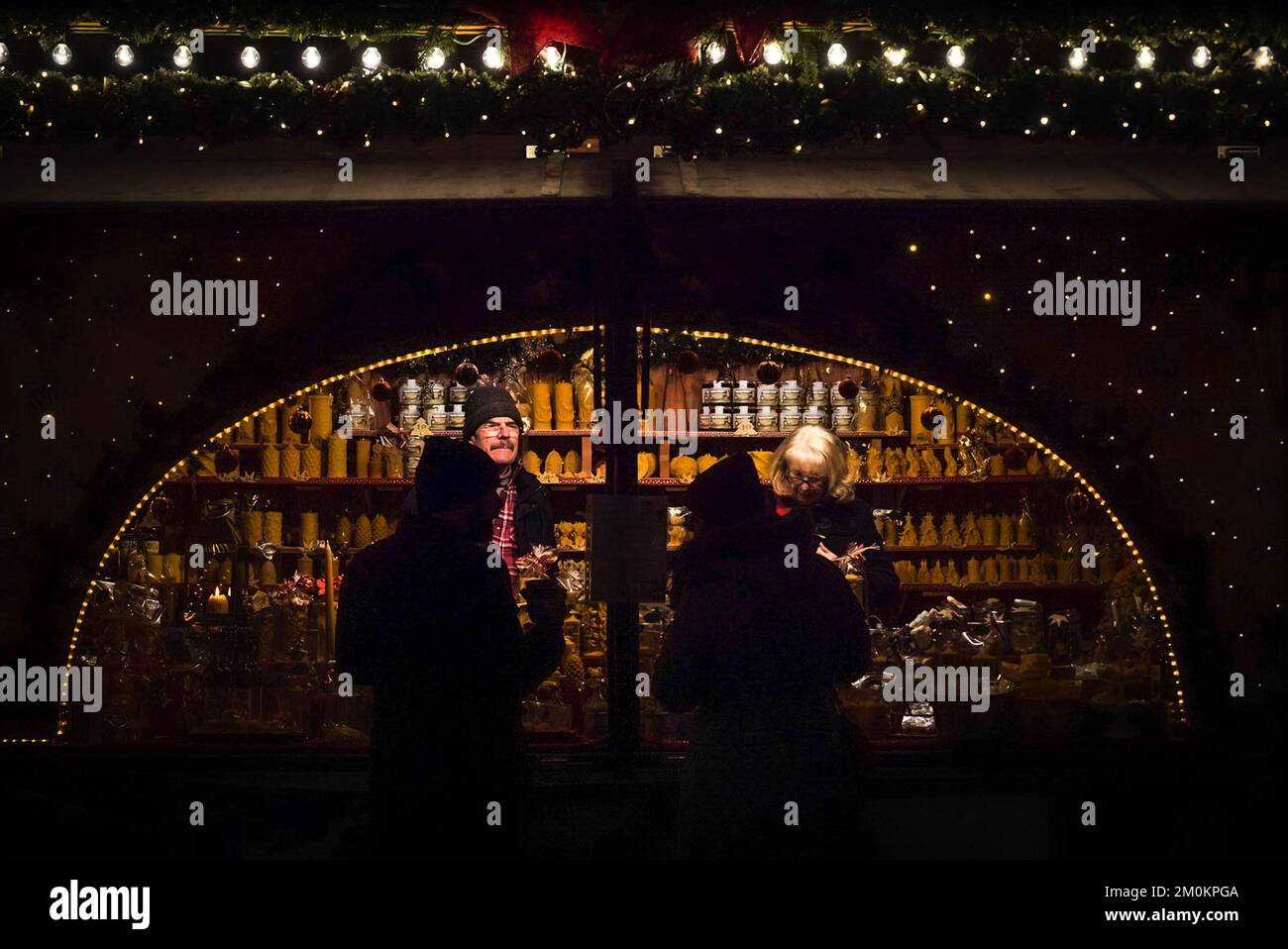 Zwei Silhouetten der Kunden stehen in der Nähe eines beleuchteten Kiosks auf einem Weihnachtsmarkt in München, Deutschland, bei Nacht mit zwei Verkäufern an der Theke. Stockfoto