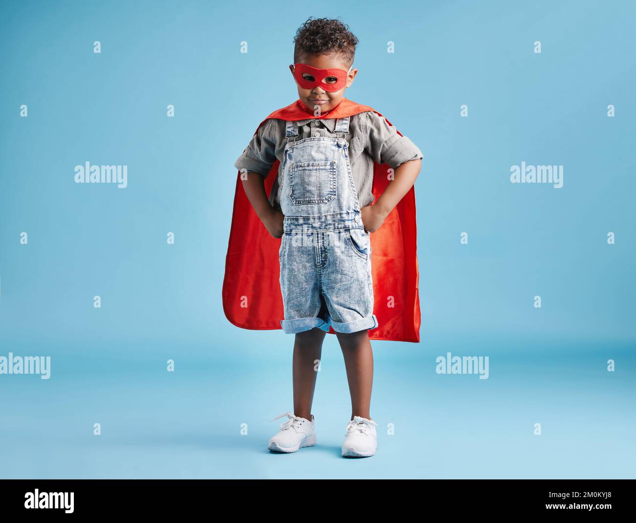 Das ganze Porträt eines tapferen kleinen Jungen in Superheldenumhang und Maske, der mit den Händen auf der Hüfte auf blauem Hintergrund steht. Ein starkes Kind, bereit, das zu retten Stockfoto