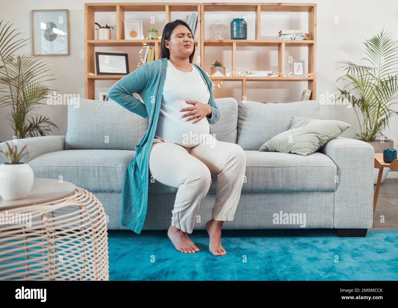 Schwangere Frau, Unbehagen und Rückenschmerzen, die auf dem Sofa im Wohnzimmer sitzen und zu Hause den Bauch halten. Frauen, die Wehen in der Schwangerschaft durchmachen, Schmerzen Stockfoto