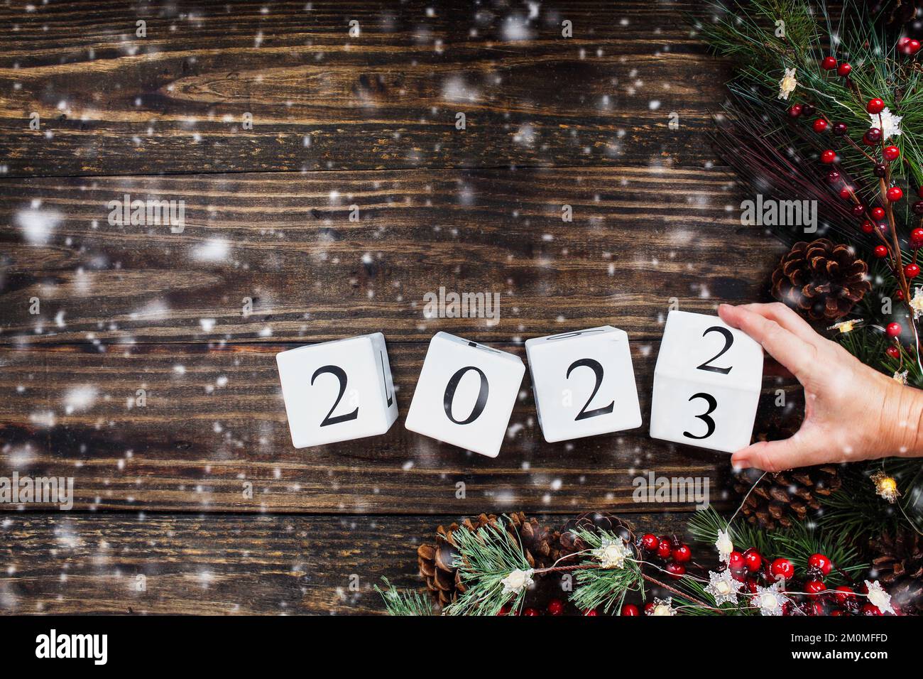 Frauenhand, die Silvester-Kalenderblöcke aus Holz für 2021 auf 2023 umwandelt. Weihnachtsbaumbeleuchtung, Kiefernäste, rote Winterbeeren und Schnee. Draufsicht. Stockfoto