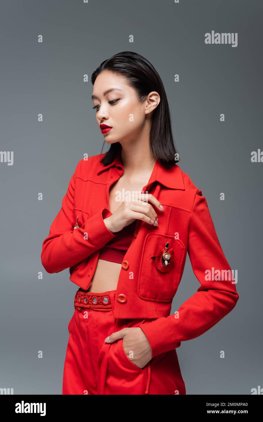 asiatische Frau in roter Hose und Jacke mit Brosche posieren mit Hand in Tasche isoliert auf grauem Stockbild Stockfoto