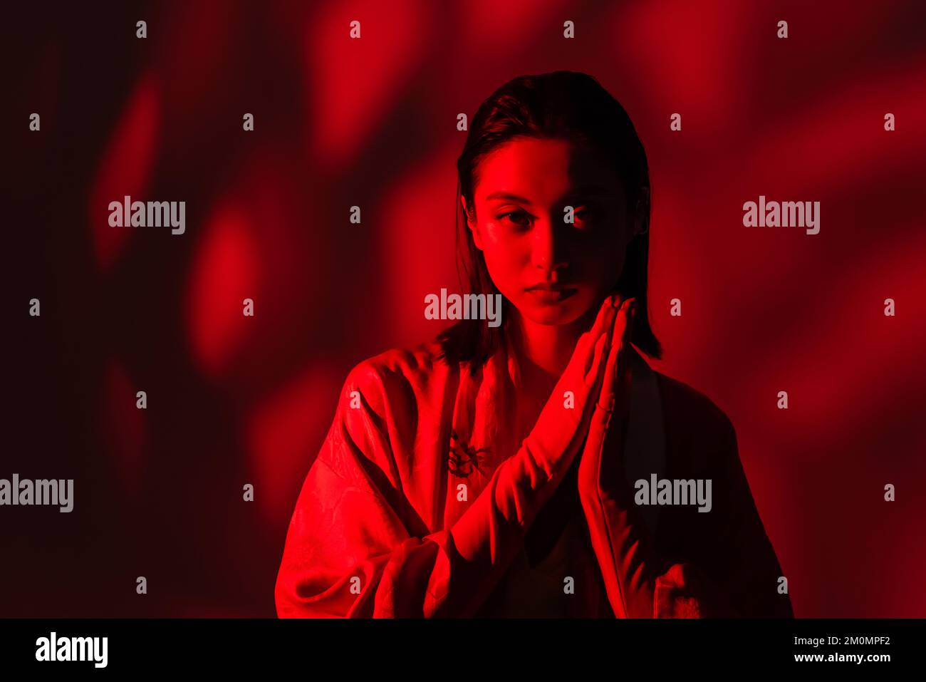 Junge asiatische Frau in Kimono-Cape mit betenden Händen auf dunklem Hintergrund mit rotem Licht, Stockbild Stockfoto