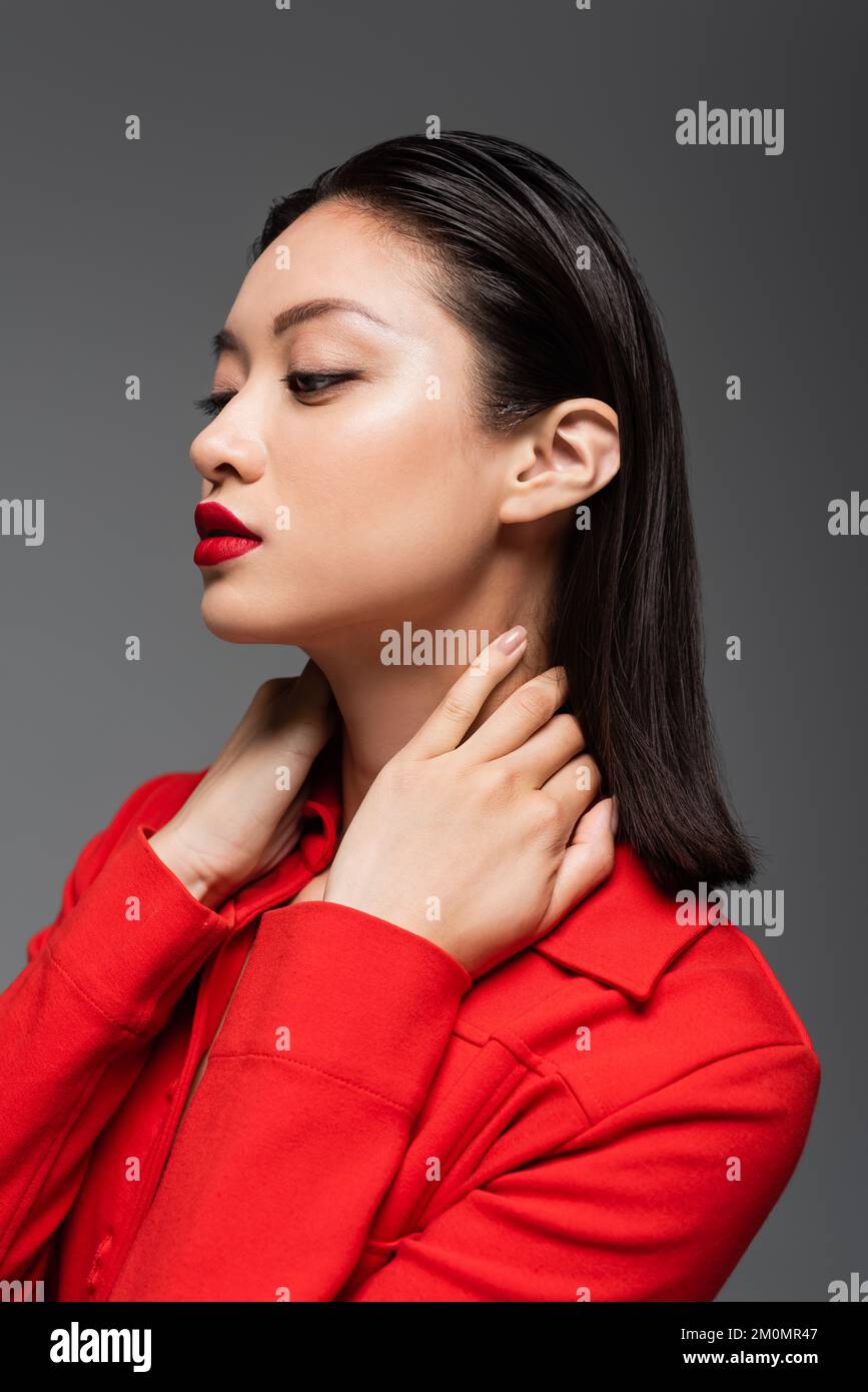 portait einer asiatischen Frau in roter Jacke mit Händen am Hals isoliert auf grauem Stockbild Stockfoto