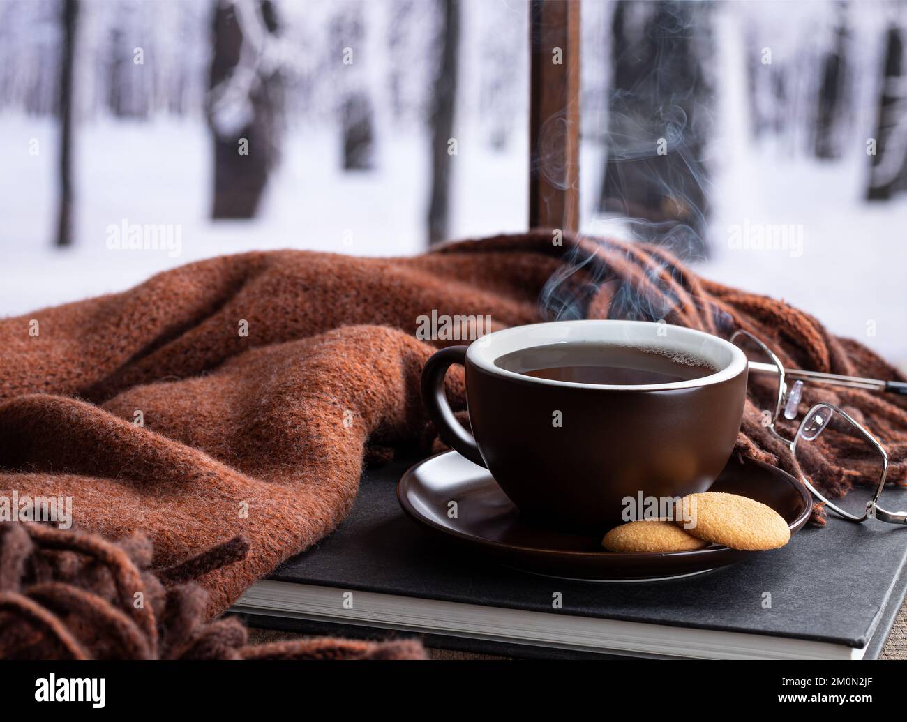 Dampfende heiße Tasse Kaffee oder Tee mit einem Buch und einer Decke an einem Fenster mit verschneitem Winterhintergrund Stockfoto