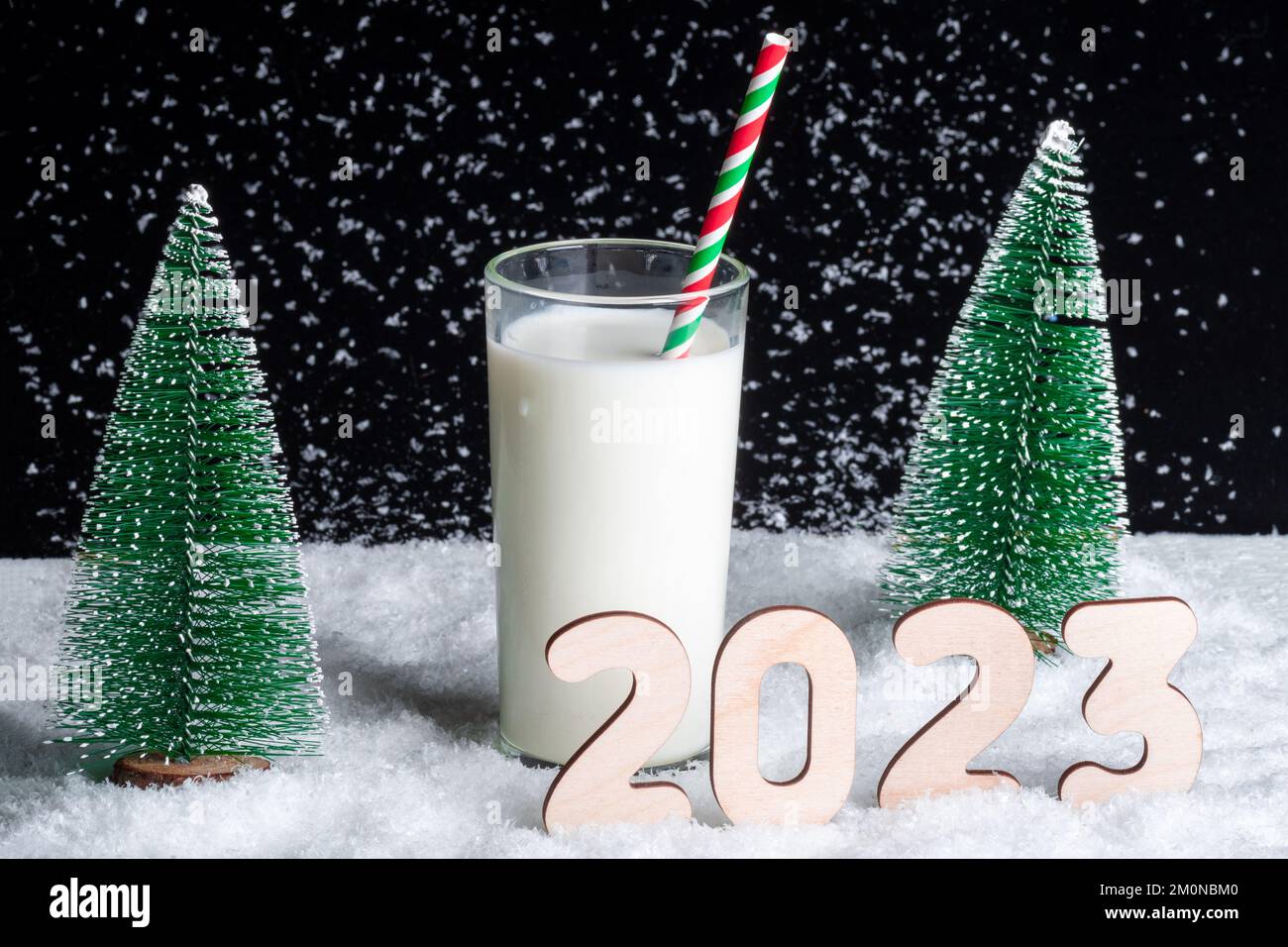 Ein Geschenk für den Weihnachtsmann. Ein Glas Milch mit Strohhalm auf verschneitem Hintergrund zwischen Weihnachtsbäumen und Holznummern 2023. Warten auf ein Wunder im N Stockfoto