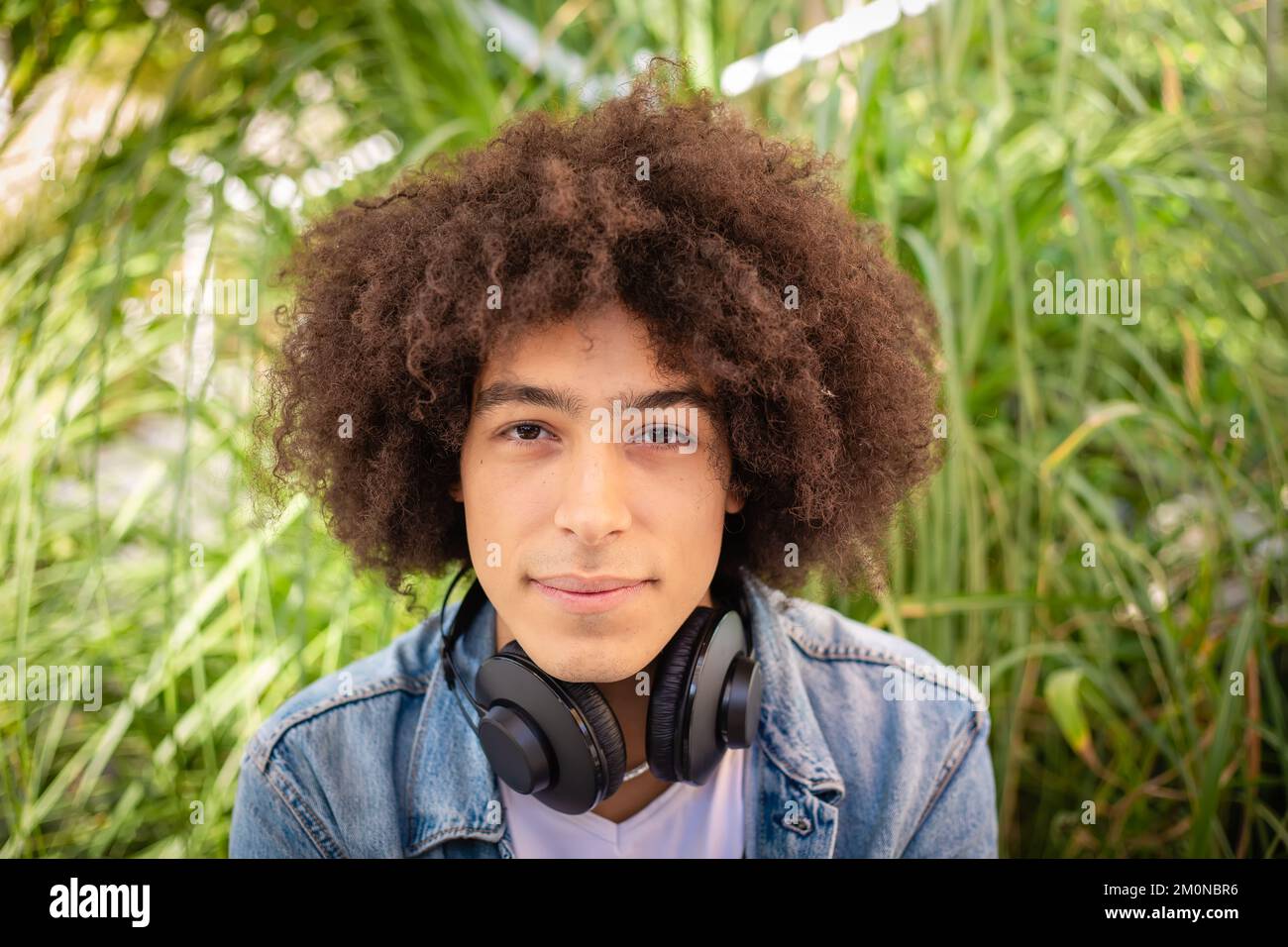 Porträt eines lächelnden jungen afro-italienischen Teenagers mit dickem lockigem Haar auf den Straßen in einem grünen Bereich mit Kopfhörern Stockfoto