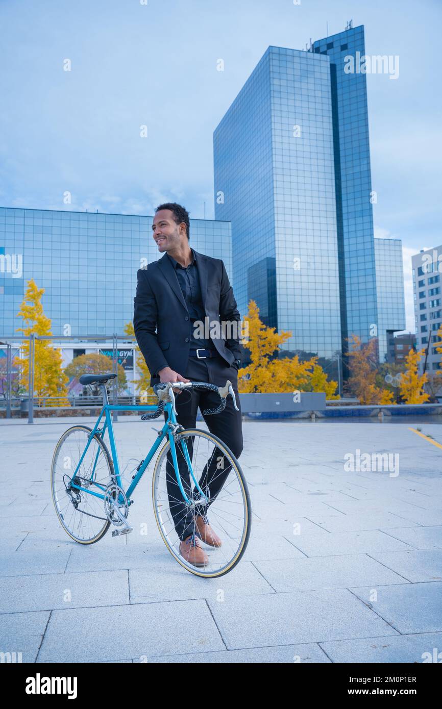 Ein junger, lächelnder, stilvoller Geschäftsmann, der mit dem Fahrrad vor dem Finanzviertel ins Büro fährt. Stockfoto