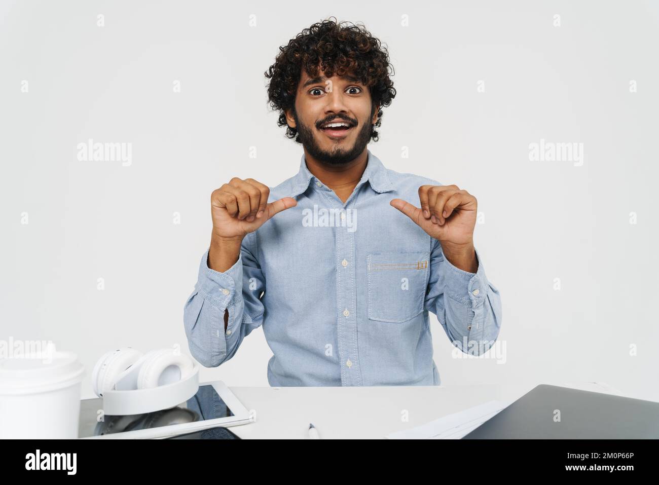 Ein erwachsener indischer Büroangestellter zeigt auf sich selbst am Bürotisch mit einem hoffnungsvollen Gesicht auf isoliertem grauen Hintergrund Stockfoto