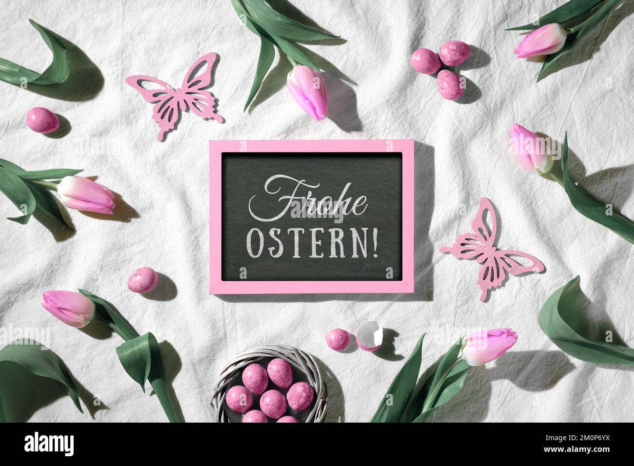 Ostern-Hintergrund. Dekorierter Hintergrund mit Tafel. Der Text Frohe Ostern bedeutet Happy Easter in deutscher Sprache. Rosa Tulpen, Holzschmetterlinge und Stockfoto